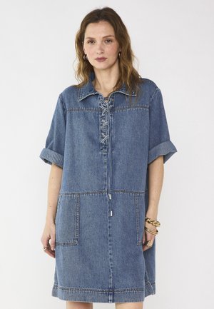 Donna che indossa un vestito in denim largo, fino al ginocchio, con chiusura frontale a lacci, maniche corte arrotolate e grandi tasche frontali, su sfondo neutro.