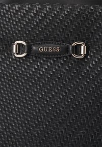 Schwarze, geflochtene Handtasche mit strukturiertem Oberflächenmaterial, Ledertrageriemen mit goldfarbener Metallakzentuierung und "GUESS"-Logo.