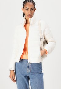 Hvit dunjakke med høy krage, kviltet design og frontlås. Båret over en oransje genser og kombinert med blå jeans.