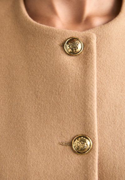Manteau en laine couleur chameau avec un col rond et deux boutons dorés ornés de motifs en relief, mettant en avant une texture lisse et douce.