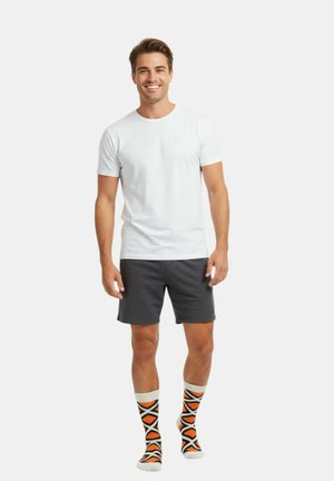 Lachende man die een effen wit T-shirt, donkergrijze korte broek en witte sokken met een oranje en zwart geometrisch patroon draagt, staand op een witte achtergrond.