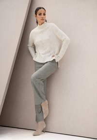 Frau, die sich gegen eine schräge Wand lehnt und einen weißen strukturierten Pullover, graue Karohosen und beiges, hochhackiges Schuhwerk trägt, in einer neutralen Umgebung.