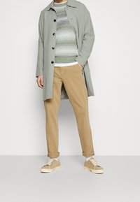Cappotto grigio chiaro con bottoni neri, maglione verde testurizzato, pantaloni beige e scarpe beige con lacci bianchi, esposti su uno sfondo semplice.