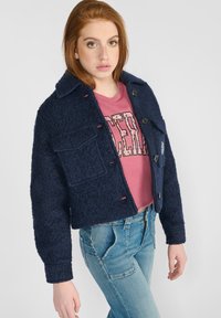 Veste cropped bleu marine en tissu texturé, avec fermeture à bouton et poche avant. Portée sur un sweat-shirt graphique rose et assortie avec un jean clair.
