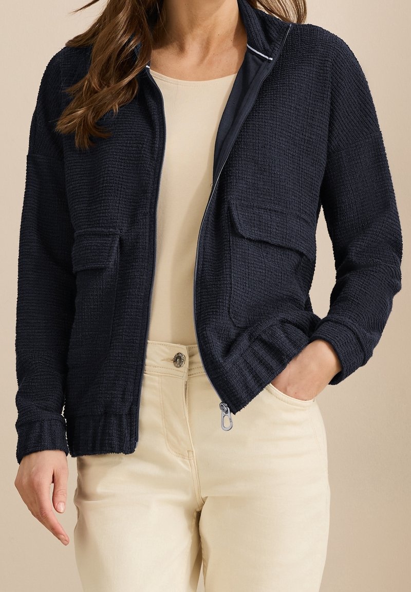 Femme portant une veste zippée bleu marine texturée avec poches à rabat sur un haut beige et un pantalon de couleur claire, debout avec une main dans la poche.
