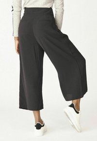 Černé široké kalhoty vyrobené z měkké, splývavé tkaniny s vysokým pasem, vybavené dvěma zadními kapsami, stylizované s bílými teniskami.