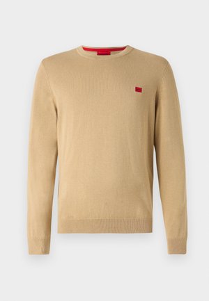 Beige genser med rund hals, lange ermer og ribbestrikkede mansjetter. Har en liten rød logo på brystet. Myk, strikket tekstur.