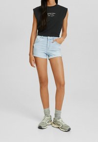 Schwarzes ärmelloses Oberteil mit bedrucktem Text, hellblaue Jeansshorts mit ausgefranstem Saum und graue Sneakers mit weißen Akzenten und dicken Sohlen.