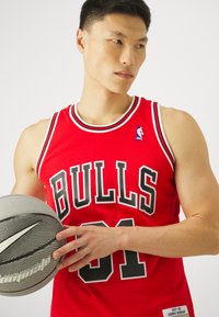 Czerwona koszulka do koszykówki z siateczkowej tkaniny, czarnymi akcentami, z dużym napisem "BULLS" i logo NBA. Trzyma szaro teksturowaną piłkę do koszykówki.