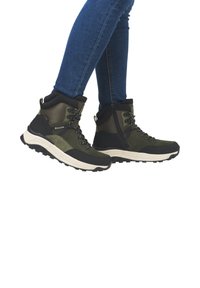 Bottes de randonnée vertes et noires avec une tige haute, en matériaux texturés, à lacets, et une semelle beige épaisse, portées avec un jean bleu.