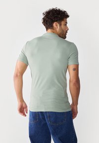 Lys grønn t-skjorte med korte ermer og tettsittende design. Har en glatt tekstur. Båret med mørkeblå denimjeans. Bakfra.