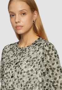 Blouse transparente grise et blanche avec un motif à pois, col rond et fermetures à boutons. Manches longues et amples avec des détails froncés.