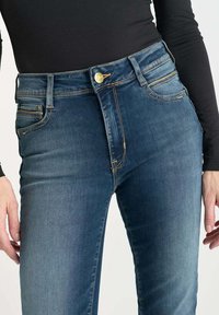 Jeans en denim bleu foncé avec une texture lisse, taille haute, design à cinq poches et accent en métal doré au bouton avant.