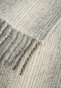 Pelēkā un krēmīgi svītrota vilnas šalle ar trauslām malām. Iespējota mīksta, teksturēta virsma un gaisīgu un tumšu šķiedru maisījums.