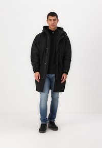 Svart parkacoat med hette, glidelås og knapper. Har en litt oversized passform og lange ermer. Brukt over en svart genser og jeans.