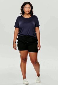 Moteris stovi vilkėdama tamsiai mėlyną marškinėlį, juodus šortus su virvele, baltas sportines batus ir apyrankes ant ausų, prieš paprastą pilką foną.