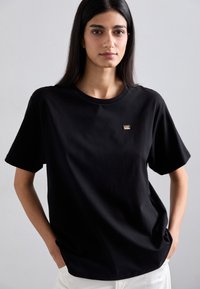 Black cotton t-shirt with short sleeves, round neckline, featuring a small gold logo accent on the chest, relaxed fit.