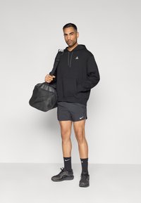 Μαύρο φούτερ με κουκούλα και μπροστινή τσέπη και λογότυπο, σκούρο γκρι σορτς, μαύρες κάλτσες και αθλητικά παπούτσια, κρατώντας μια μαύρη τσάντα duffel, ουδέτερο φόντο.