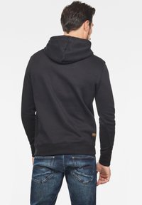 Svart huva sweatshirt med en texturerad finish, med ribbad nederkant och ärmar. Bärs med blå jeans, med en gul etikett på sidan.