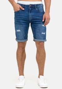 Shorts en denim bleu moyen avec des détails délavés. Jambes retroussées et fermeture à bouton. Associés à des baskets blanches.