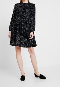 Robe noire à pois blancs, manches longues, encolure arrondie et taille élastique. Portée avec des chaussures plates noires.