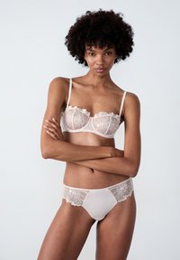 Koronkowy biustonosz i pasujące majtki w delikatnym beżu z kwiatowym haftem, subtelnymi ramiączkami i przezroczystymi elementami. Gładka faktura z falbaniastymi krawędziami.