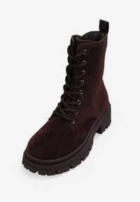 Botines de ante marrón hasta el tobillo con puntera redonda, diseño de cordones, ojales de metal y suela de goma gruesa con un patrón de textura en la banda de rodadura.