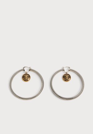 Par de pendientes de aro en tono plateado con rayas negras que presentan pequeños colgantes dorados con cabeza de Medusa colgando dentro de cada aro sobre un fondo blanco.