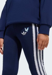 Tmavomodré legíny s bielymi riasenými bočnými pruhmi, s motýľovým dizajnom a logom Adidas na stehne. Vyrobené z elastickej látky.