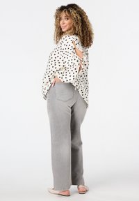 Femme aux cheveux bouclés portant un blouse blanche à cœurs noirs et un jean gris, posant avec une main dans la poche, regardant par-dessus son épaule.