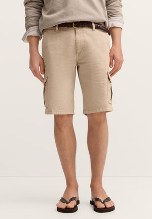 Person wearing beige cargo shorts, brown braided belt, light sweater, and black flip-flops standing against a plain background.