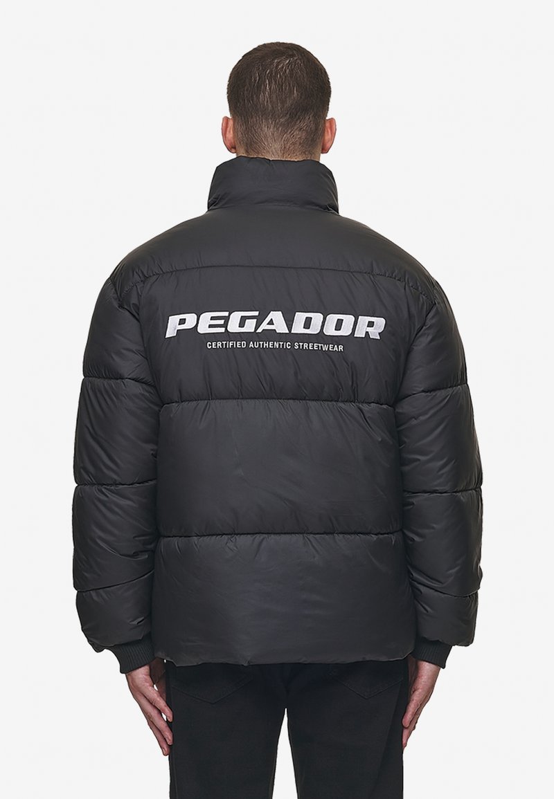 Muž v černé péřové bundě s vysokým límcem, na zadní straně s bílým nápisem "PEGADOR Certified Authentic Streetwear", stojící zády k nám.