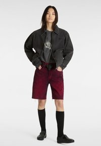 Chaqueta de mezclilla negra sobre una camiseta gráfica gris, combinada con pantalones cortos desgastados rojos y calcetines negros. El calzado presenta tachuelas y un diseño simple.