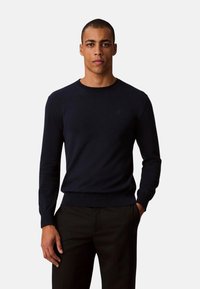 Jersey de punto azul marino con cuello redondo y puños acanalados, con un logo sutil en el pecho. Combinado con pantalones oscuros.