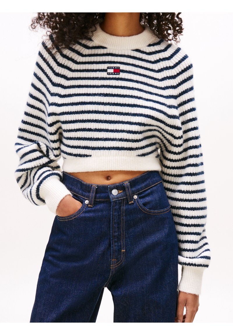 Femme portant un pull court blanc avec des rayures horizontales bleu marine et un jean taille haute bleu foncé, main dans la poche, torse visible.