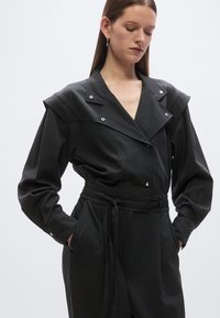 Črni kombinezon z velikim ovratnikom, dolgimi rokavi in pasom s sponko. Ponaša se z zaskočni zapenjalci in stranskimi žepi, izdelan je iz gladkega blaga.