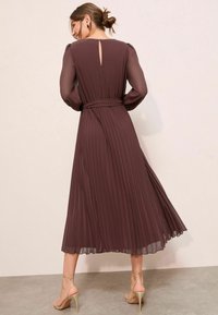 Langes, plissiertes Kleid in einem satten Mauveton. Mit langen Ärmeln, einem Schlüsselloch-Ausschnitt im Rücken und einem Taillengürtel. Getragen mit beigen, hochhackigen Sandalen.
