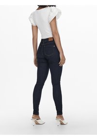 Mujer con una blusa blanca de mangas abullonadas, vaqueros ajustados azul oscuro con un parche de cuero, y mules blancos de tacón alto, de pie de espaldas.