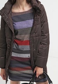 Manteau bouffant marron avec une capuche, doté de quincaillerie argentée. En dessous, une robe rayée rouge, grise et violette avec un léger éclat.