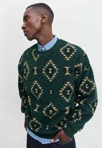 Pull en polaire vert présentant des motifs en losanges et en triangles beige et noir. Manches et ourlet côtelés, superposé sur une chemise col bleu clair.