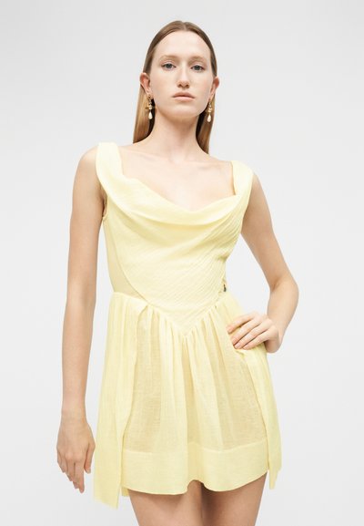 Νέα γυναίκα με ίσια ξανθά μαλλιά, που φοράει αμάνικο απαλό κίτρινο φόρεμα με ντραπέ λαιμόκοψη και χρυσά σκουλαρίκια κρεμαστά.