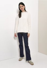 Pull en maille côtelée blanc, jeans évasés bleu foncé avec coutures blanches, et baskets gris clair. Design simple, look décontracté.