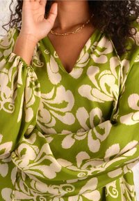 Top in tessuto verde con motivi floreali astratti bianchi, scollatura a V e maniche ampie con polsini arricciati. Accento con collana in catena dorata.