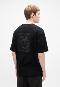 Joven con cabello corto castaño claro mirando hacia la derecha, vistiendo una camiseta negra con un patrón abstracto texturizado en la parte superior de la espalda.