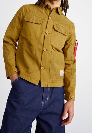 Mann trägt eine senfgelbe, geknöpfte Jacke mit vier Fronttaschen und einem roten Etikett am Ärmel, kombiniert mit dunkelblauen Hosen.