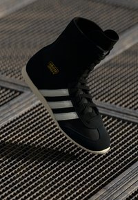 Zapatilla alta negra con diseño de tres franjas blancas, parte superior de tejido texturizado, suela de goma y detalles en dorado en el costado.