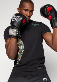 Chemise noire à manches courtes avec un motif camouflage sur le côté, affichant le texte vert "Venum". Le mannequin porte des gants de boxe noirs et rouges.