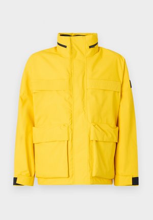 Kollane veekindel jakk kõrge krae, esine tõmblukk ja kolm suurt taskut. Omab musti detaile randmete ja krae lähedal.