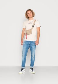 Beige stof crossover tas med flere rum og en rød lynlås, båret over en lys t-shirt og blå jeans, på en neutral baggrund.
