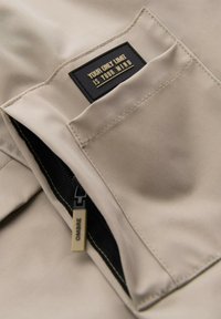 Beiger Stoff mit einer Tasche, die einen schwarzen Reißverschluss und ein Patch mit der Aufschrift "DEINE EINZIGE GRÖSSE IST DEINEN GEIST" aufweist. Reißverschluss-Zipper mit der Bezeichnung "OMBRE".